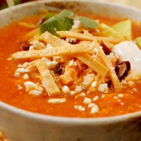 Bowl of red colored Mexican sopa de lima, with tortillas, and other ingredients in the middle of the bowl.