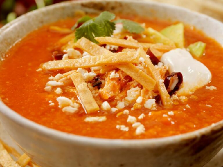 Bowl of red colored Mexican sopa de lima, with tortillas, and other ingredients in the middle of the bowl.