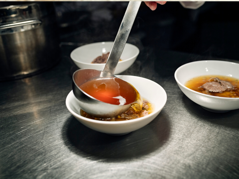 Ladle pouring consomme over meat in a small white bowl