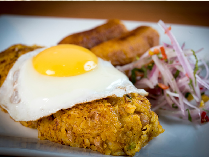 White plate with tacu tacu and a fried egg with a side of sausages and onions.